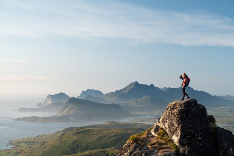 Person with backpack on stands on a peak overlooking a grand vista. A light blue sky with subtle clouds and mountains are seen in the background.