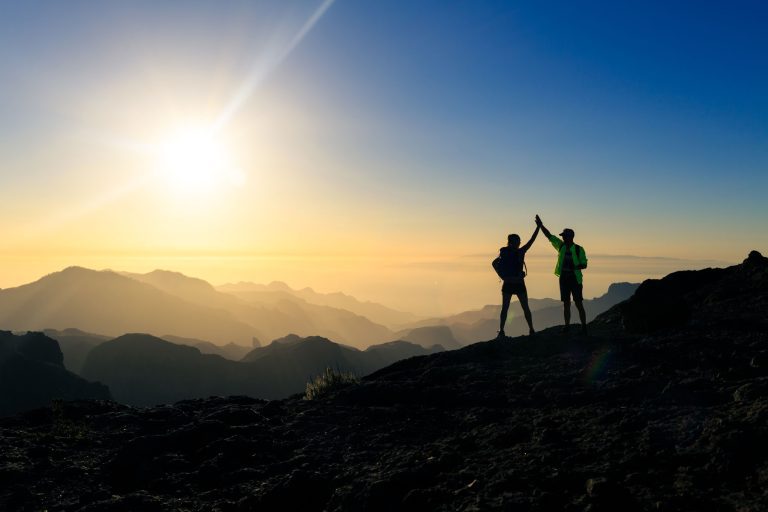 Two hikers high five each other standing on top of a mountain. We see the sun in the background as well as numerous other mountain peaks.