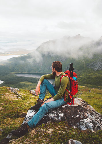 A man with a backpack, jeans and a green long sleeve shirt sits on a mossy rock looking away from the camera. We see clouds and mountains in the background.