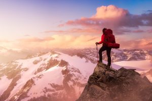 A solo hiker stands on a mountain peak facing the horizon. In the background we see snow covered mountains and several clouds. The sun is off camera and appears low in the sky.