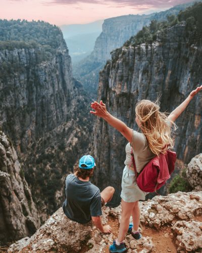 shutterstock_21506621372 A couple overlooks a massive canyon. The male is wearing a blue hat and grey shirt, facing away from the camera sitting on the cliffs edge. The blond haired woman with a red knapsack stands behind him facing the canyon with her arms spread wide.