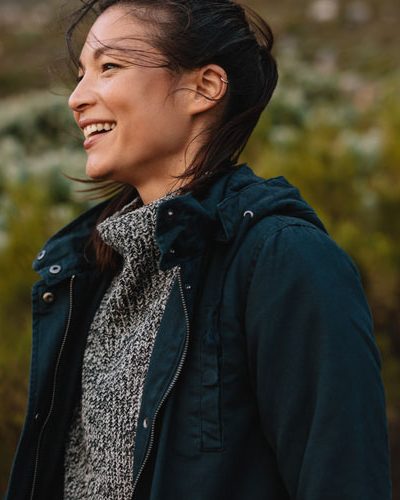 young woman smiling with a breeze at her back, wearing a deep green jacket and a grey sweater